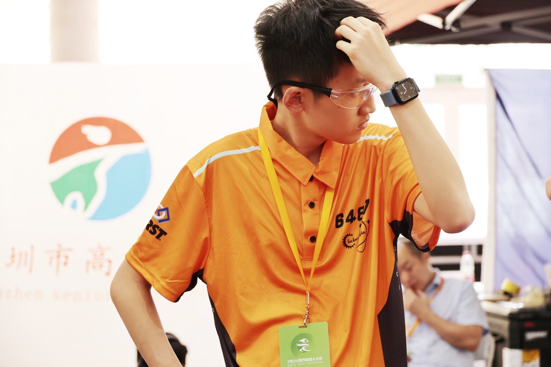 Jackson in safety glasses and an orange team shirt concentrating during a FIRST Robotics competition