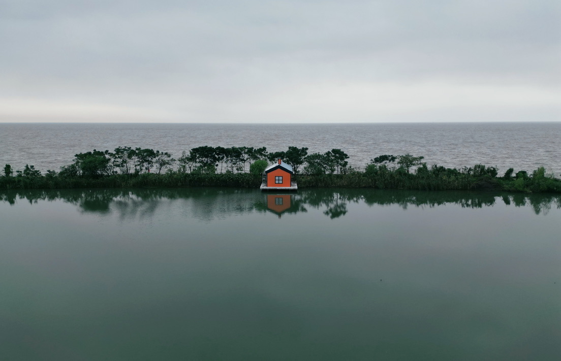 An orange cabin on a narrow spit of trees, reflected in the still water of a lake at dusk